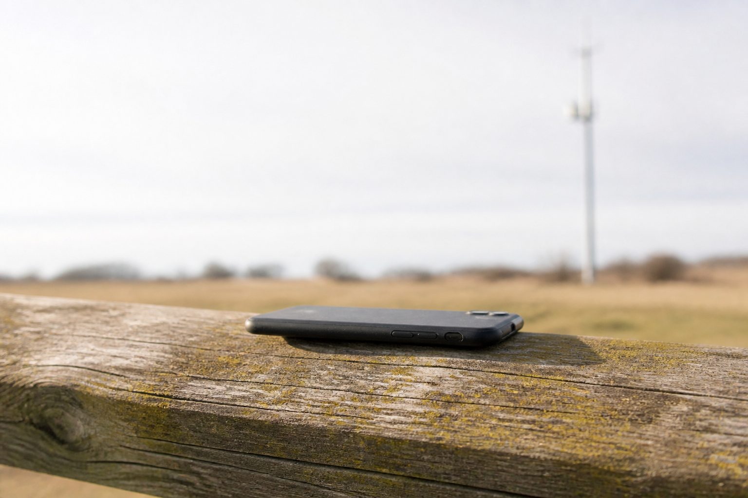 A dark unmarked phone on a weathered wooden rail, a mast out of focus across a pale field, no logo.