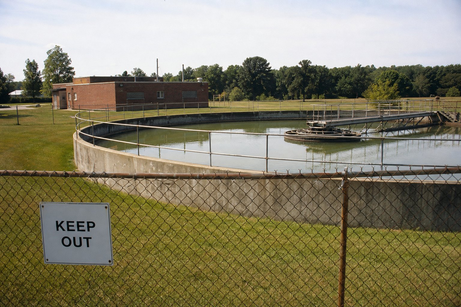 A clarifier tank behind a chain-link fence and a blank KEEP OUT sign.